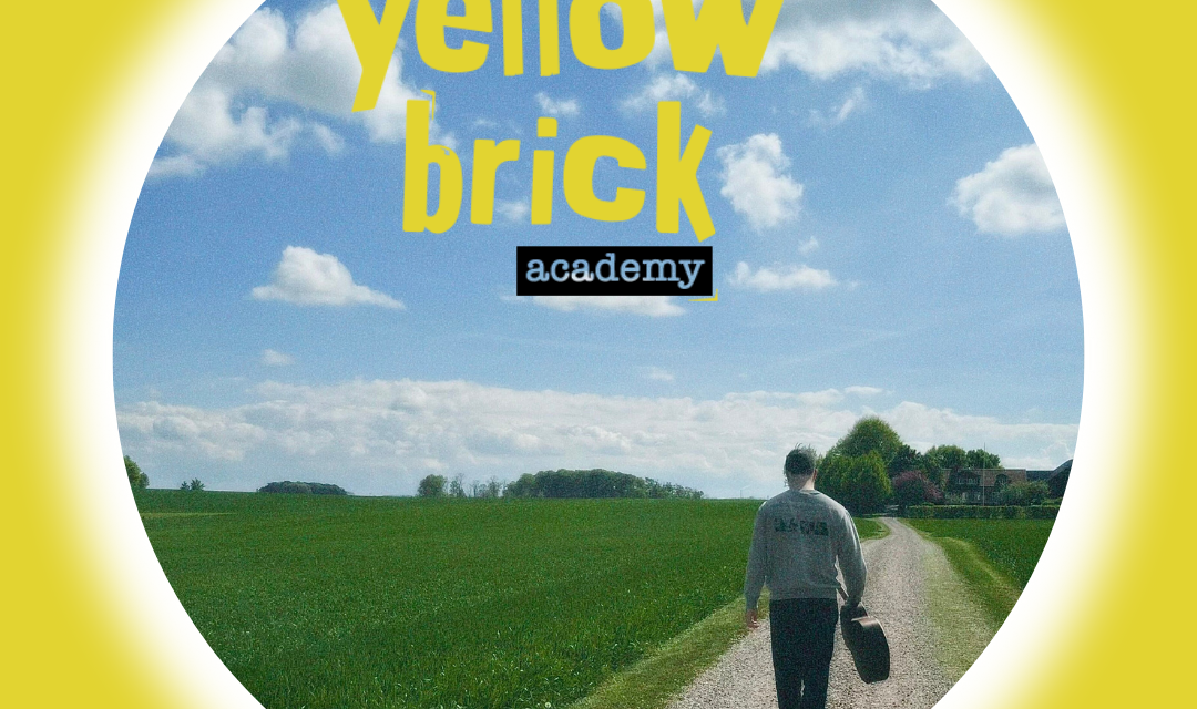 En person går bort från kameran på en grusväg i en solig dag omgiven av gröna fält. Yellow Brick Academys logga syns i bland molnen.
